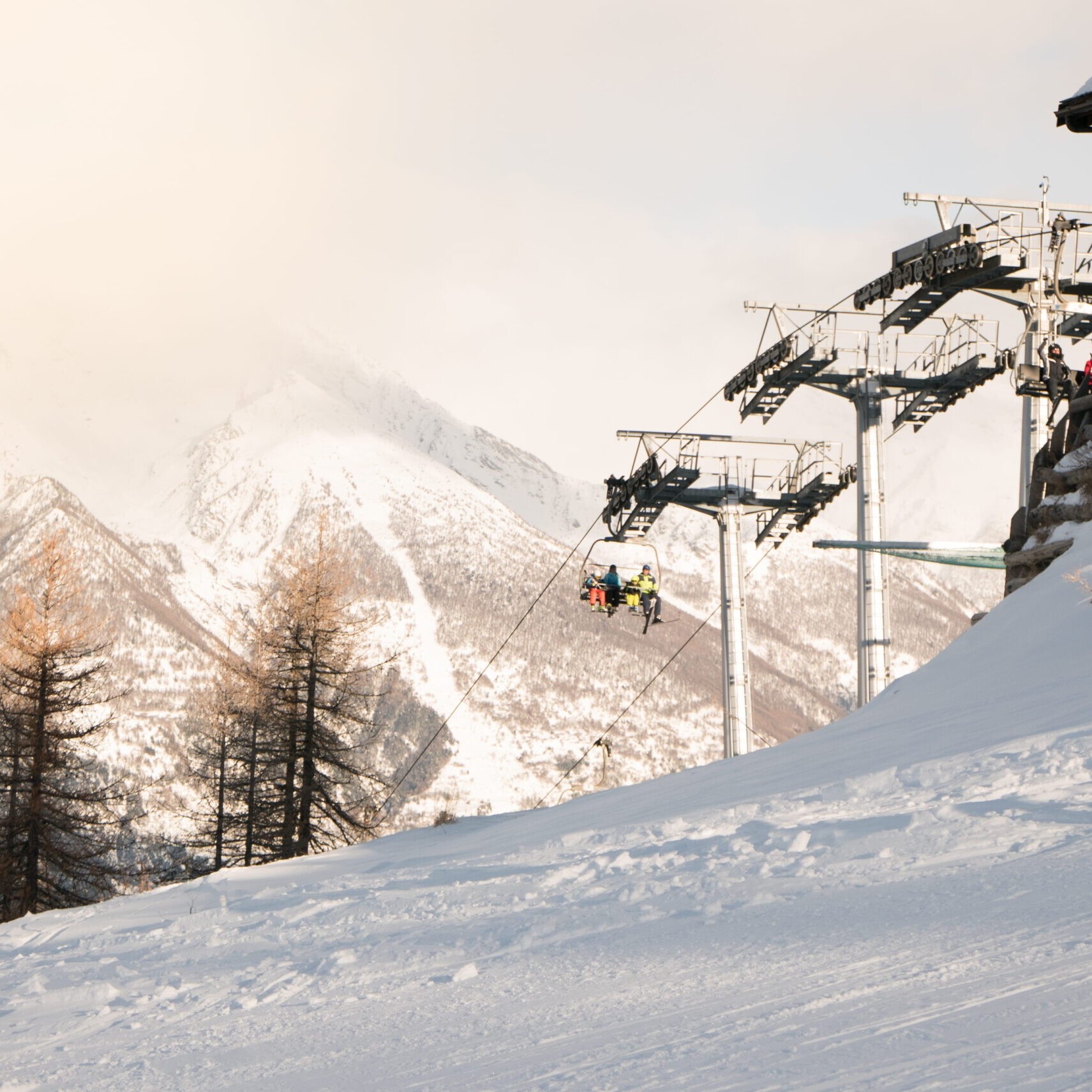 Télésiège transportant des skieurs au-dessus d’une piste enneigée dans une station de ski avec chalet et montagnes alpines en arrière-plan.