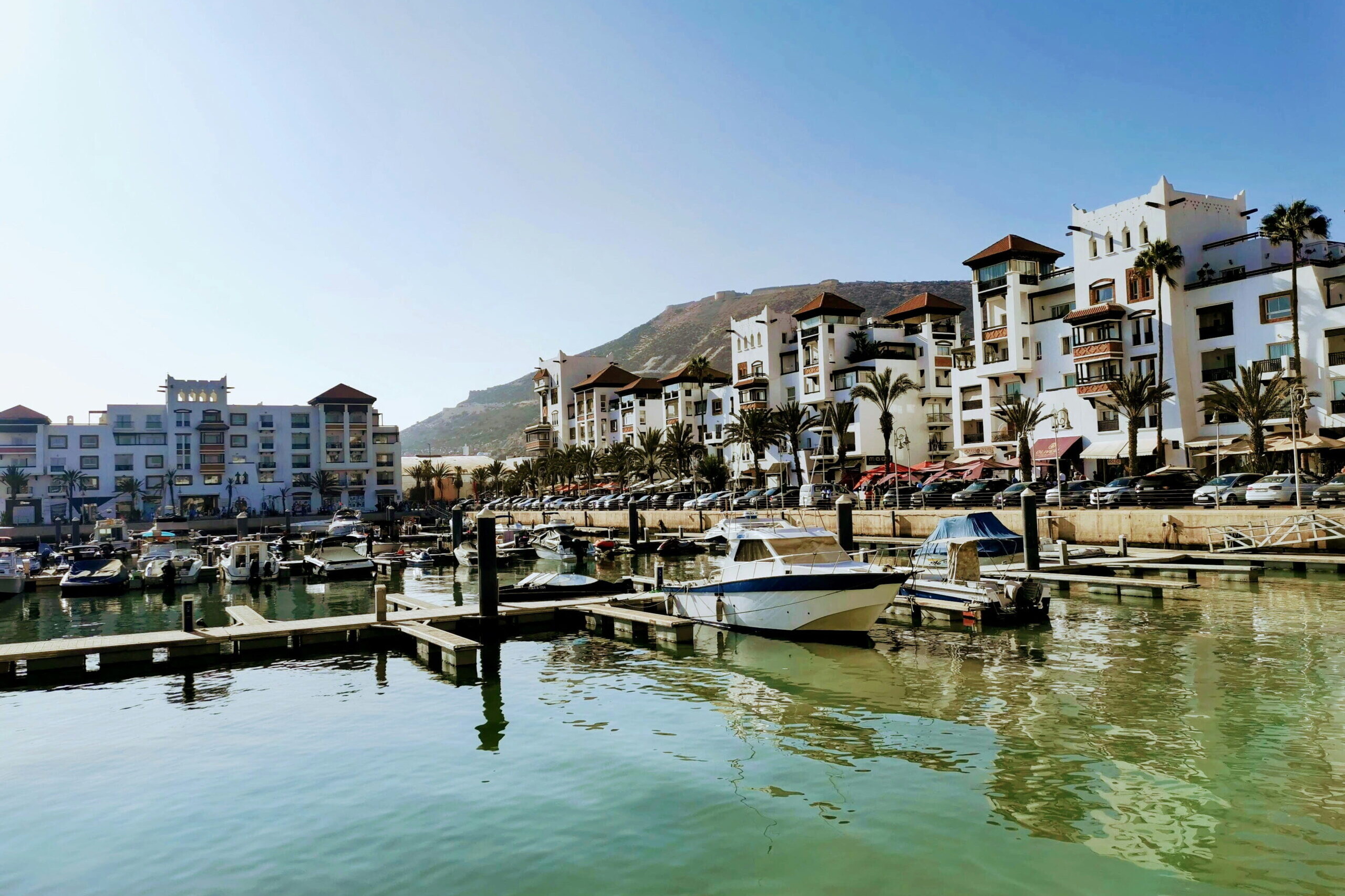 Marina d&rsquo;Agadir avec des bateaux amarr&eacute;s, des immeubles blancs bord&eacute;s de palmiers et les montagnes en arri&egrave;re-plan sous un ciel bleu.