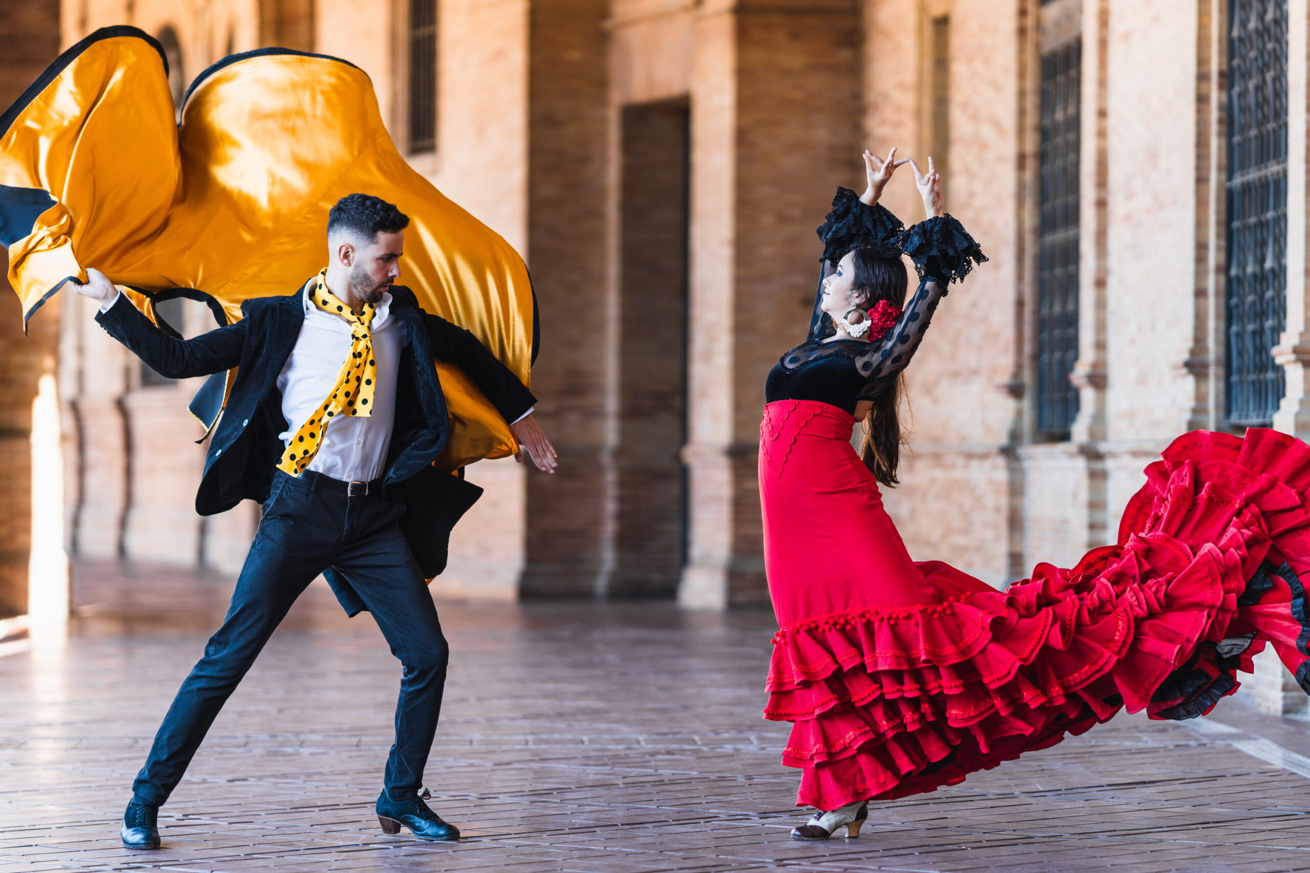 Couple de danseurs ex&eacute;cutant un flamenco &agrave; Madrid, femme en robe rouge et homme tenant une cape dor&eacute;e sous une galerie monumentale.