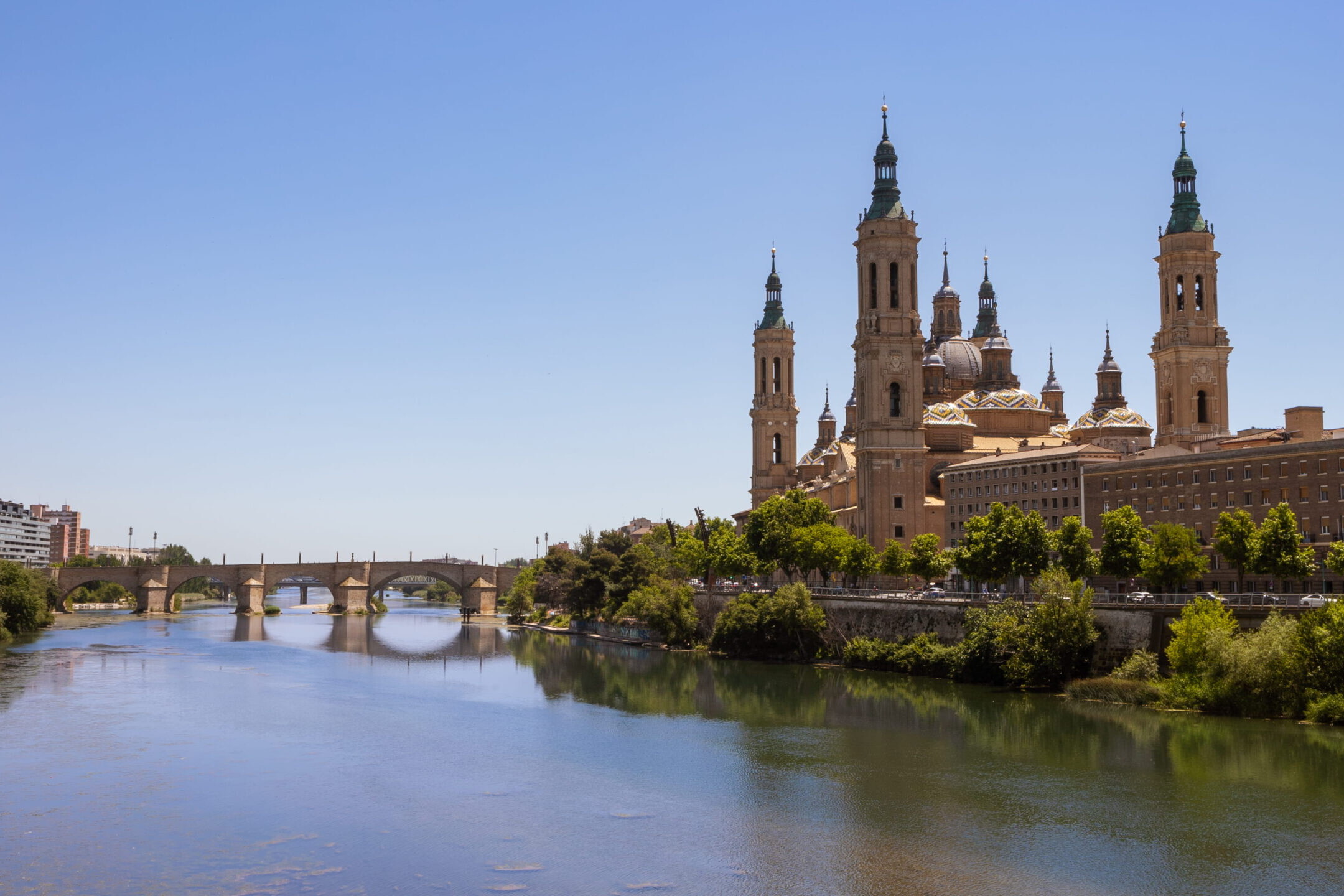 Basilique Notre-Dame du Pilar à Saragosse vue depuis le fleuve Èbre avec un pont historique et les tours de la cathédrale sous un ciel bleu.